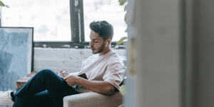 A man sat on a sofa looking down towards a notebook he's holding. 