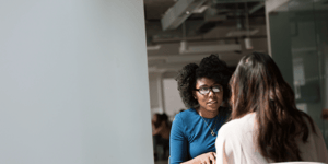 Two women talking face to face at an office desk.