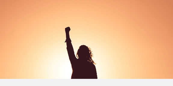 A silhouette of a woman with her fist up looking towards the sunset.