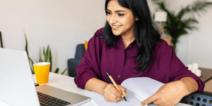 Young woman sat on a desk writing on a notepad looking at a Macbook.
