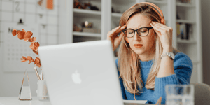 A stressed woman with her eyes closed and hands to her head facing her laptop.