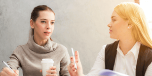 Two business women sat side by side looking towards each other while talking.