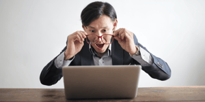A man looking shocked at his laptop screen holding his glasses in front of his face.