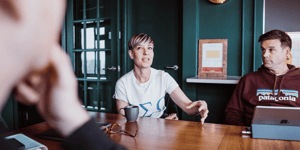 A woman and a man sat around a table. The woman is in the middle of speaking to a man not pictured.