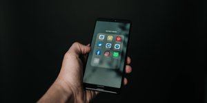 Person (not pictured) holding an iPhone. Screen shows a folder of apps called social media which includes apps like Twitter, Facebook, Snapchat, Whatsapp, and more.