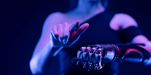 A woman with one hand pointing to the camera and her other hand, robotic and made out of metal, is held out in front of her with a closed fist.