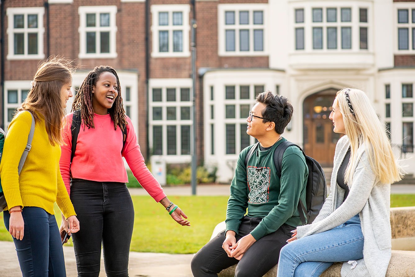 A group of 4 students chat outside Loughborough University's red brick campus building