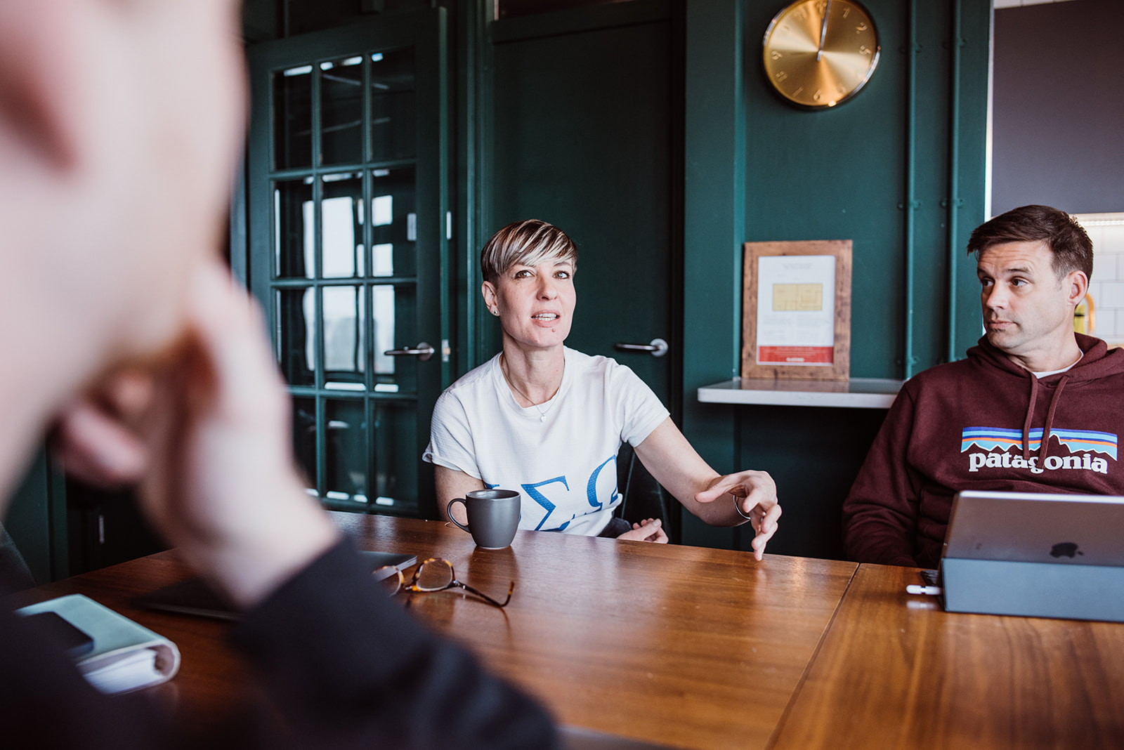 People are sat around a large wooden table, in the middle a woman in a t-shirt is talking, using her hands to help express herself